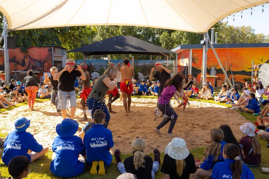 Dancers performing on the sand of the Corroboree grounds in daylight while school students watch from the grass.