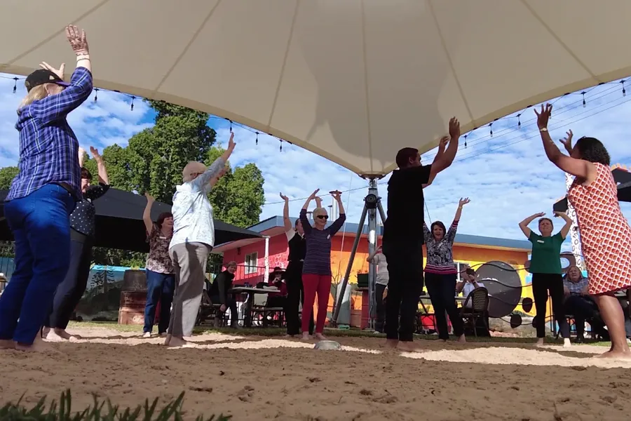 Guests with arms raised, dancing on the sand of the Corroboree grounds beneath the shade structure.