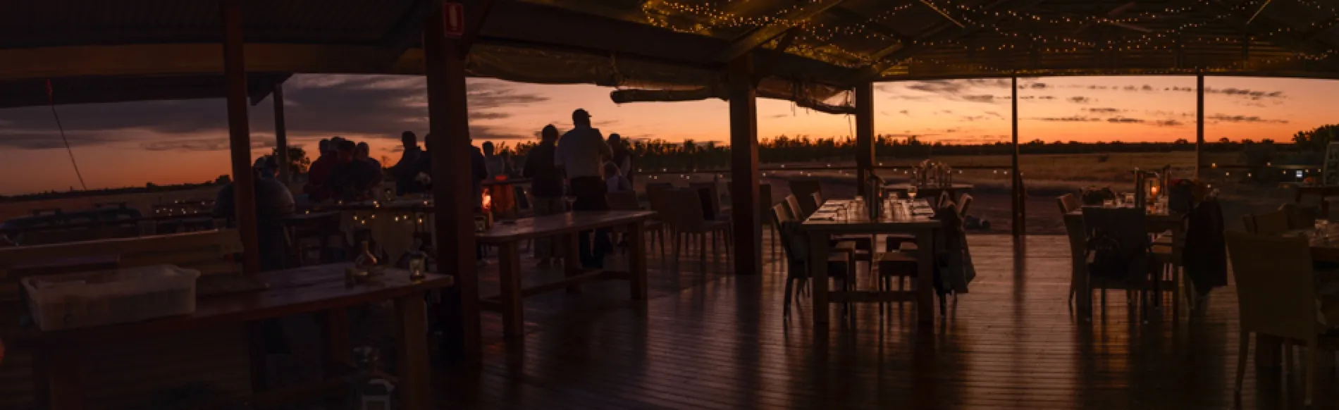 Long dinner tables set under festoon lights on an outdoor deck at dusk, looking out over open plains.